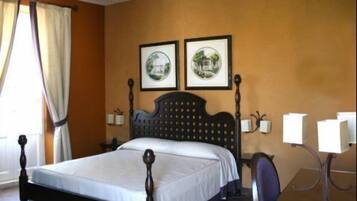 A hotel room with a wooden four-poster bed, two framed pictures on the wall, a desk with a chair, and a lamp.
