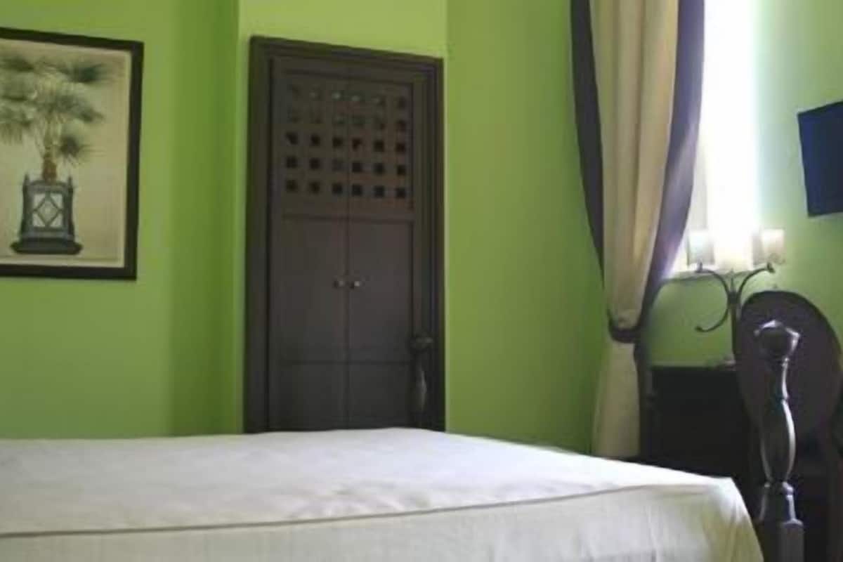 Ein Zimmer mit einem Bett, einer Tür, einem gerahmten Bild, einem Tisch mit einer Vase und einem Fenster mit Vorhängen.