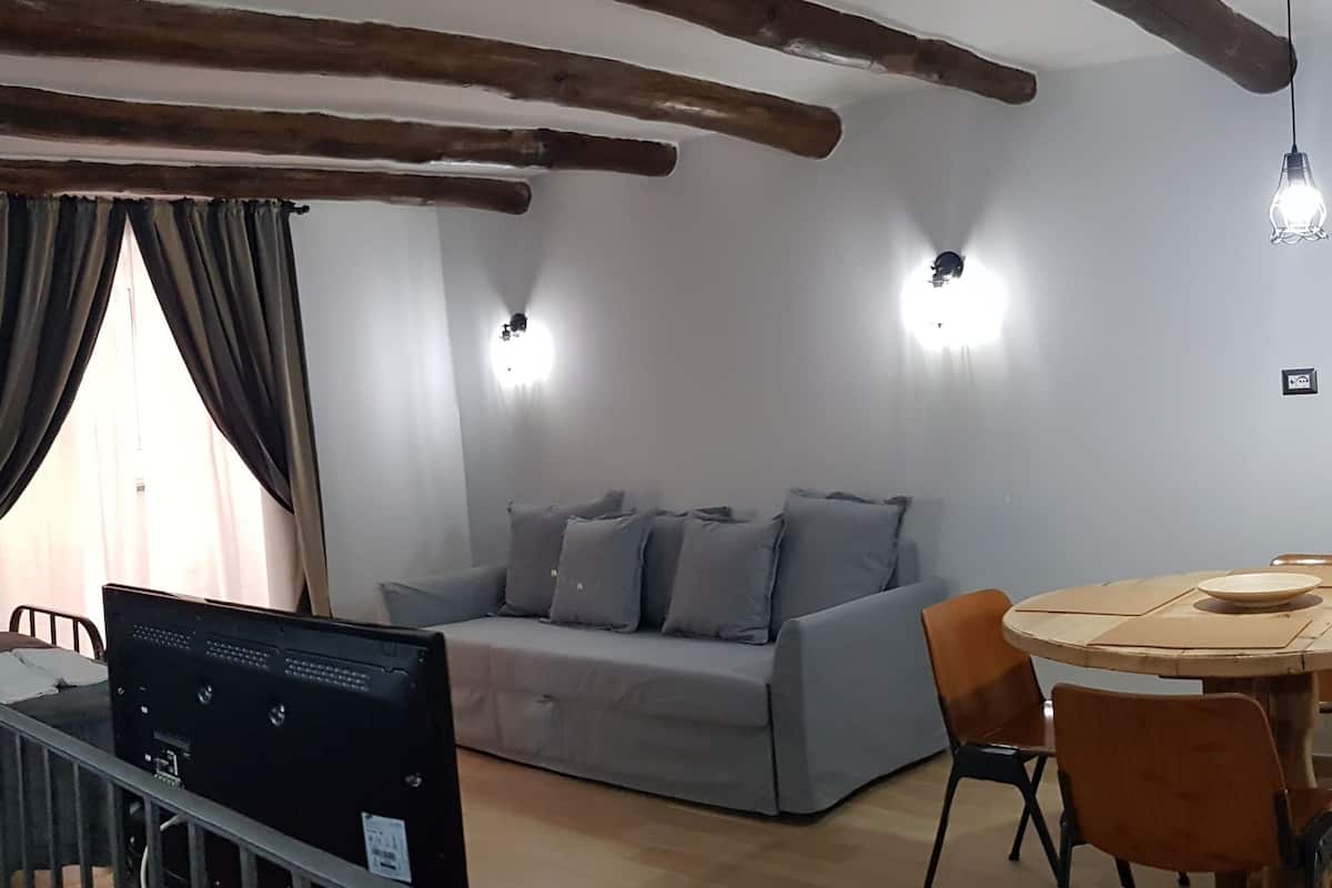A room with a grey sofa, a wooden dining table with chairs, a laptop on a desk, and a window with curtains.