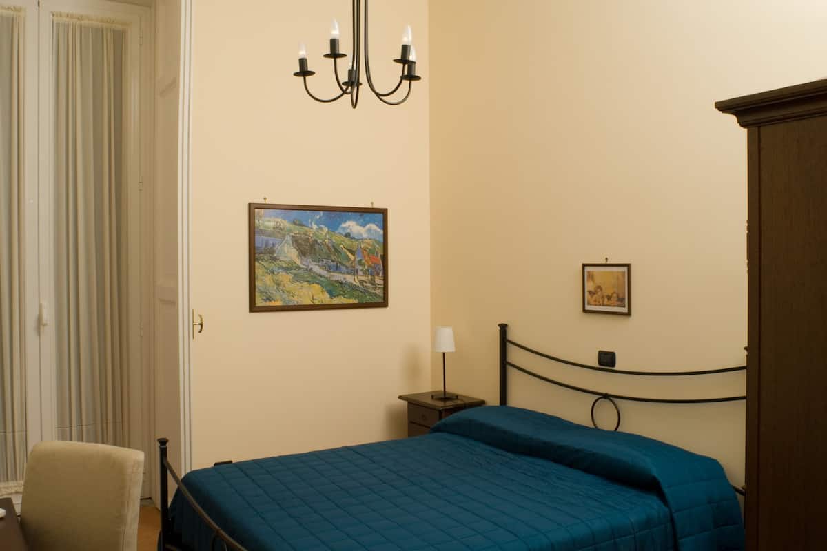 A bedroom with a bed, a chair, a nightstand, a lamp, and two paintings on the wall.