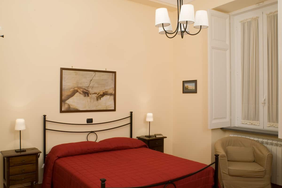 A bedroom with a bed, a nightstand, a lamp, a chair, a painting, and a window with curtains.