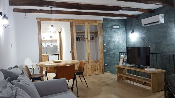 A cozy living room with a wooden ceiling, a dining area with a round table and chairs, a television on a wooden stand, and a large window. - Portanova (Naples)