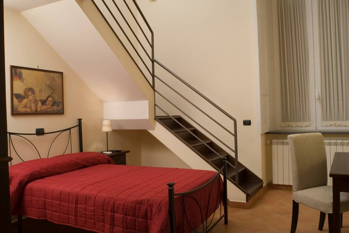 A bedroom with a bed, a nightstand, a lamp, a chair, a staircase, and a window with blinds.