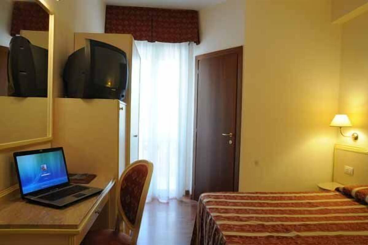 A hotel room with a bed, a desk with a laptop, a chair, a television mounted on the wall, and a door.