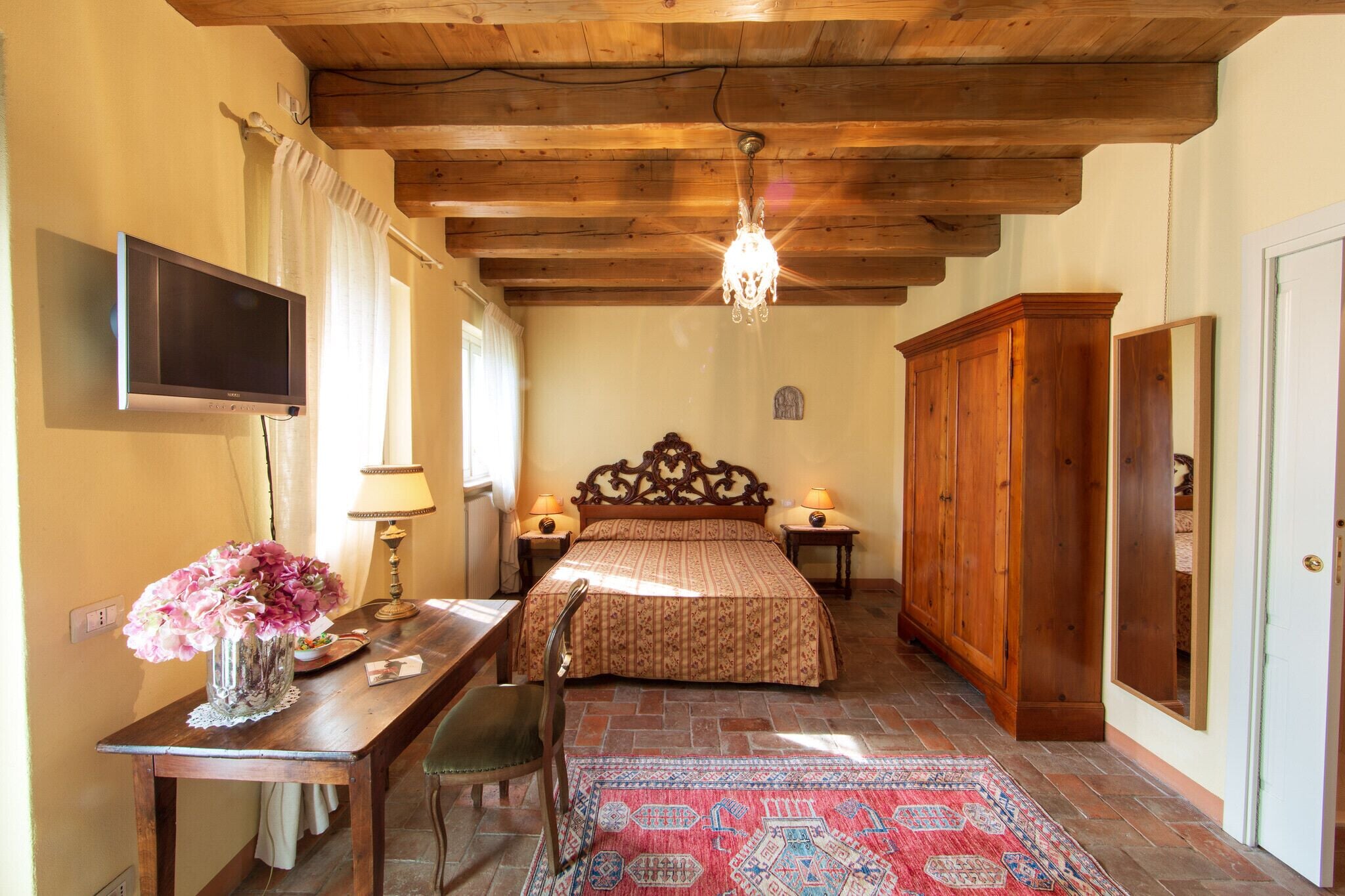 Une chambre avec un plafond en bois, un lit recouvert d’une couvre-lit à motifs, une table de chevet avec une lampe, une armoire en bois, un téléviseur fixé au mur et un vase à fleurs posé sur une table.