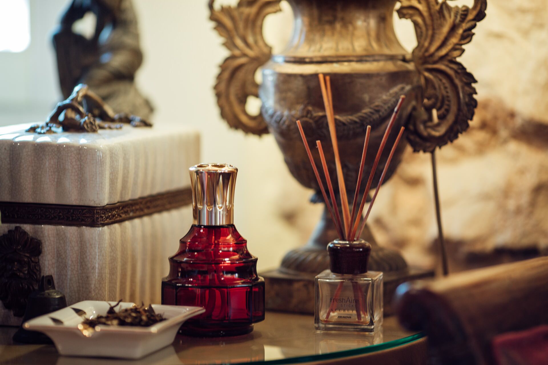 A table with a red incense burner, a small glass bottle with a label, and a white dish with what appears to be tea leaves.
