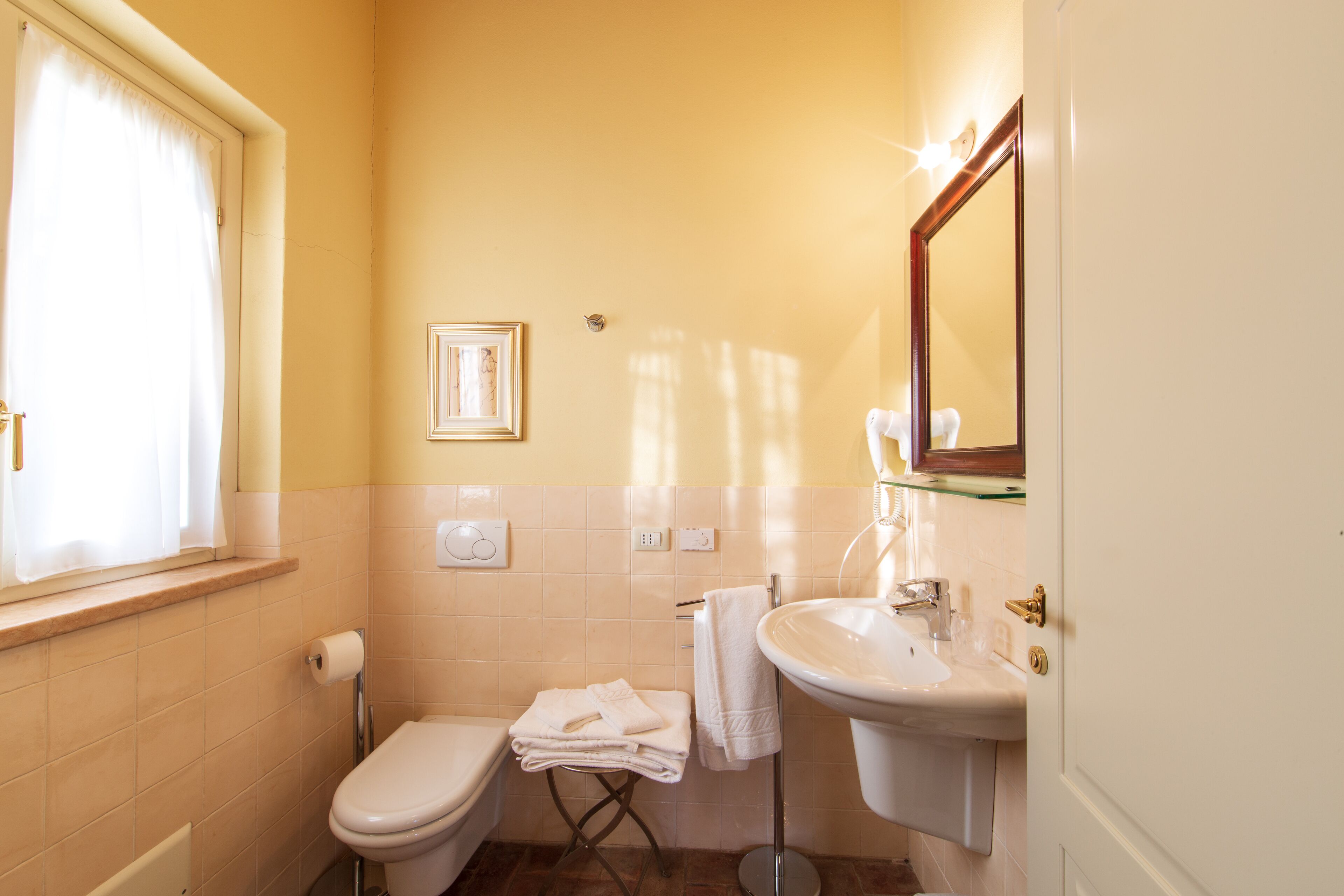 A bathroom with a toilet, sink, and mirror. There is a window with curtains, a towel rack, and a framed picture on the wall.