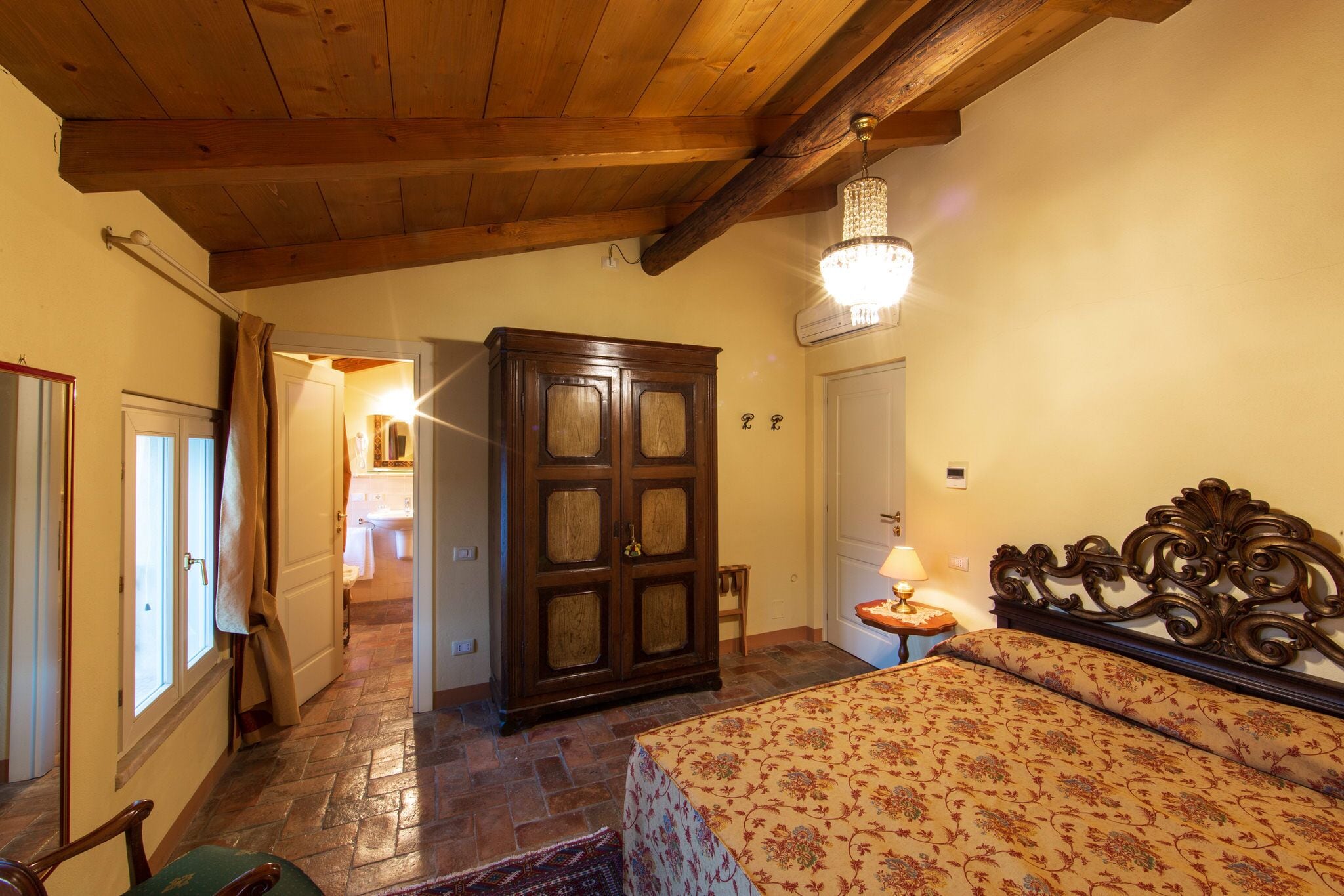 A bedroom with a wooden ceiling, a chandelier, a bed with a floral bedspread, a wooden wardrobe, and a small bedside table with a lamp.
