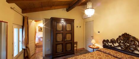 A bedroom with a wooden ceiling, a chandelier, a bed with a floral bedspread, a wooden wardrobe, and a small bedside table with a lamp.