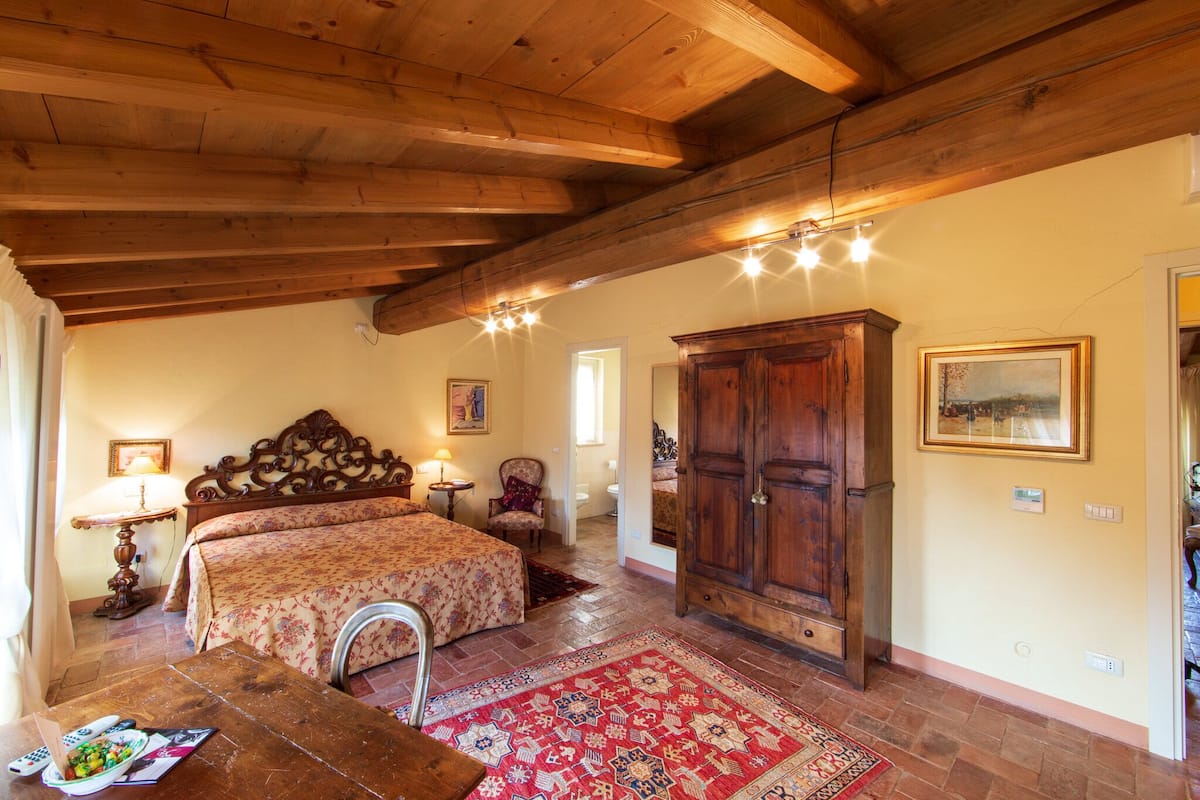 Une chambre avec un plafond en bois, un grand lit, une armoire, un bureau avec une lampe et un tapis rouge à motifs.