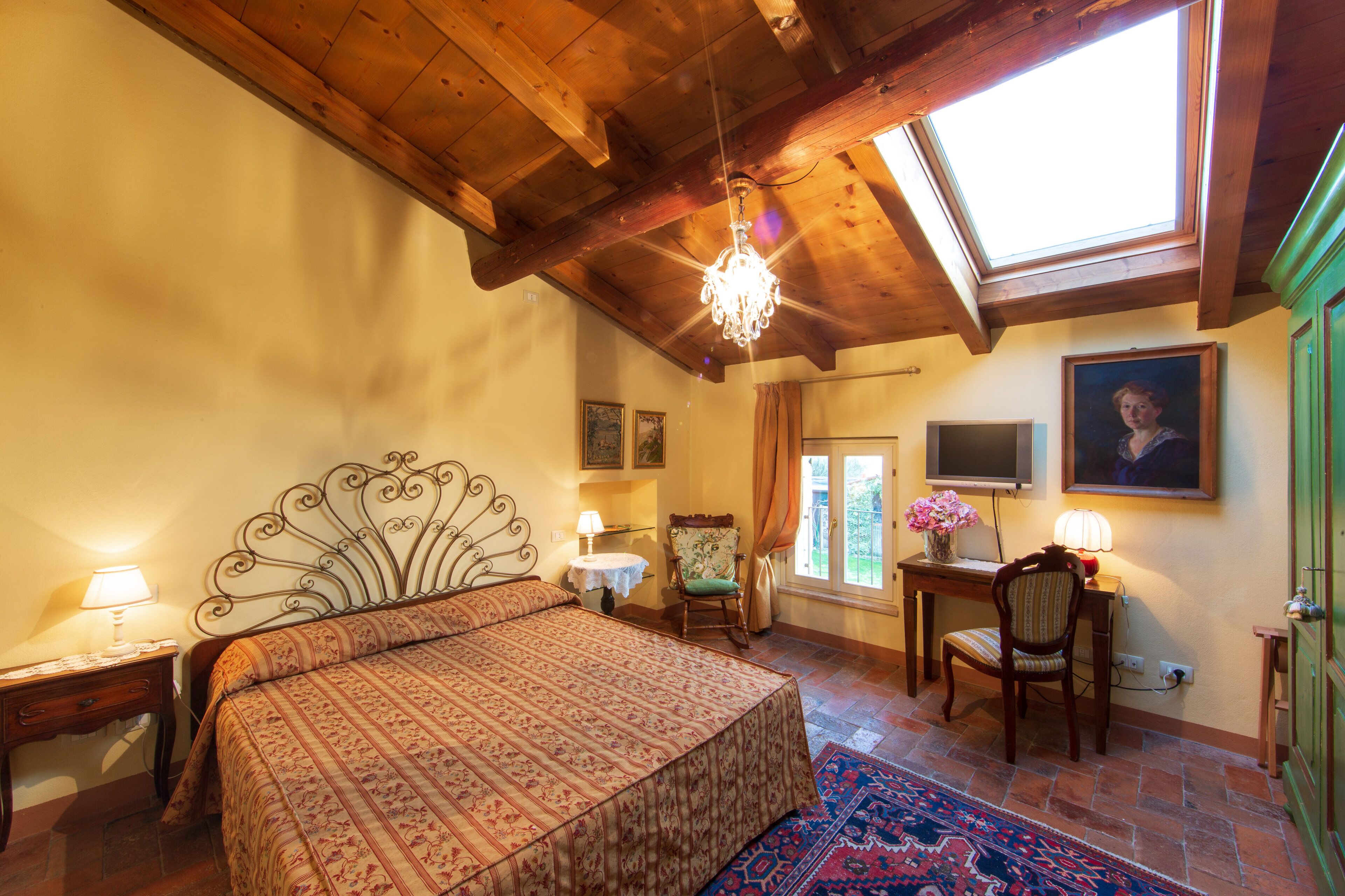 A bedroom with a large bed, a desk, a chair, a TV, and a painting on the wall.
