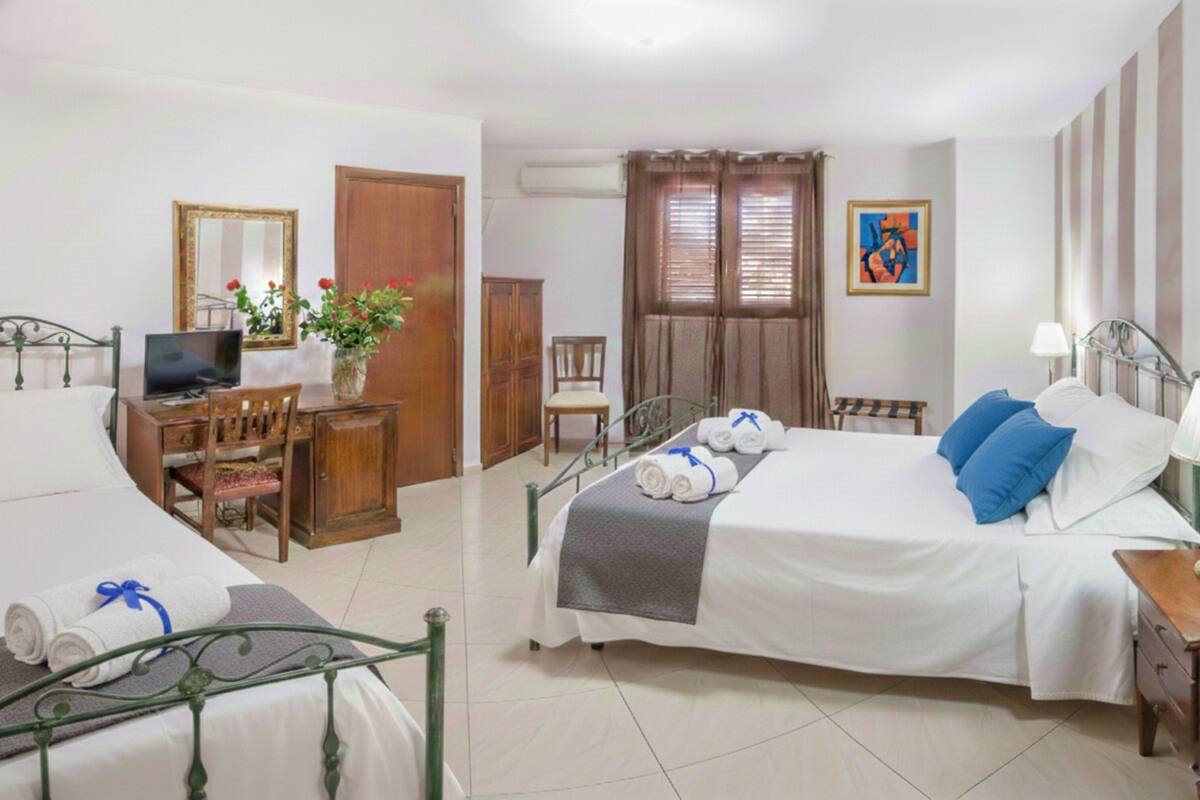 A hotel room with two beds, a desk with a computer, a chair, a TV, a plant, and a painting on the wall.