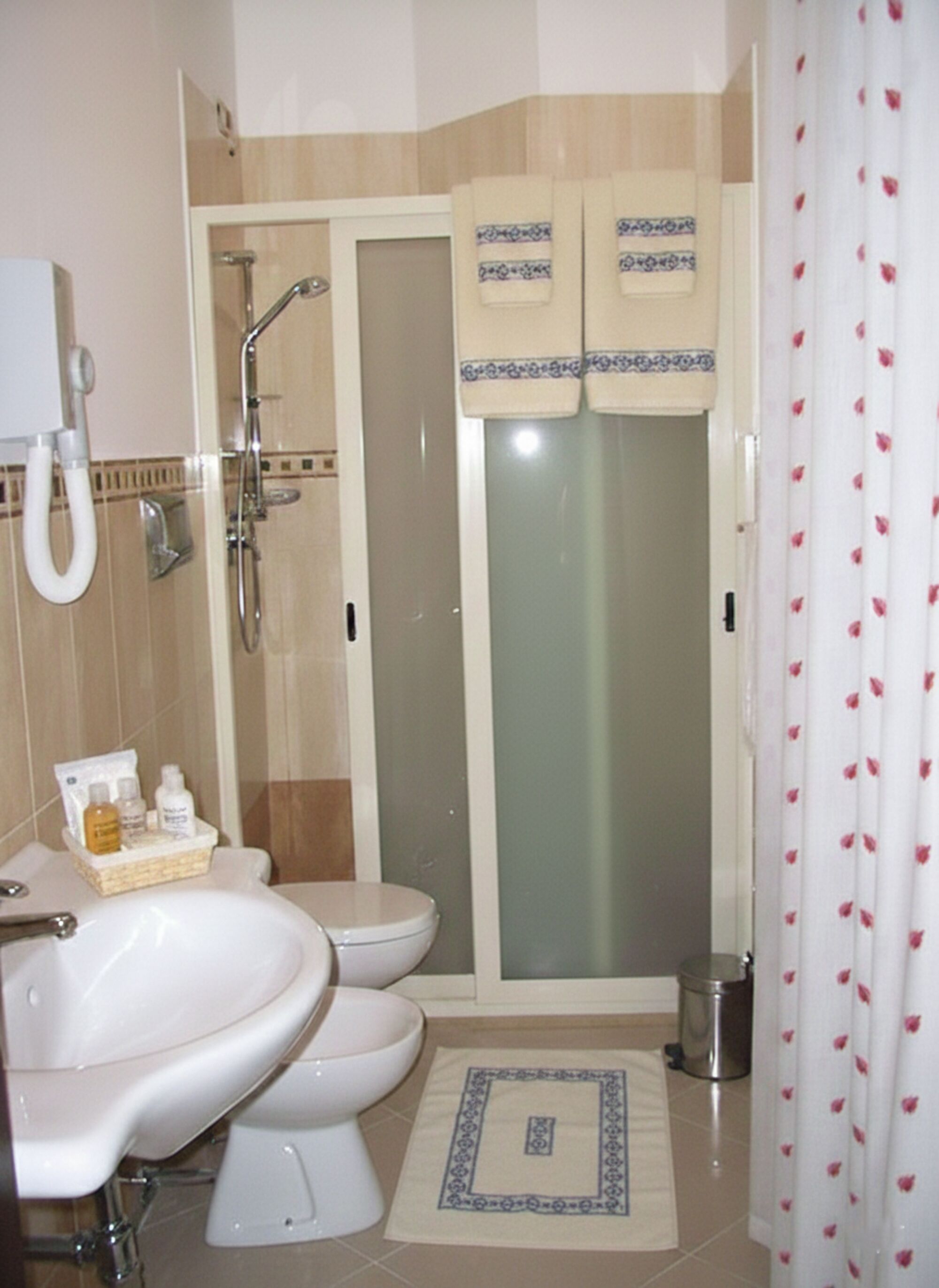 A bathroom with a sink, two toilets, a shower, and a trash can.