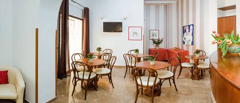 Un espace repas avec des tables rondes et des chaises, un téléviseur fixé au mur et une table recouverte d’une nappe rouge.