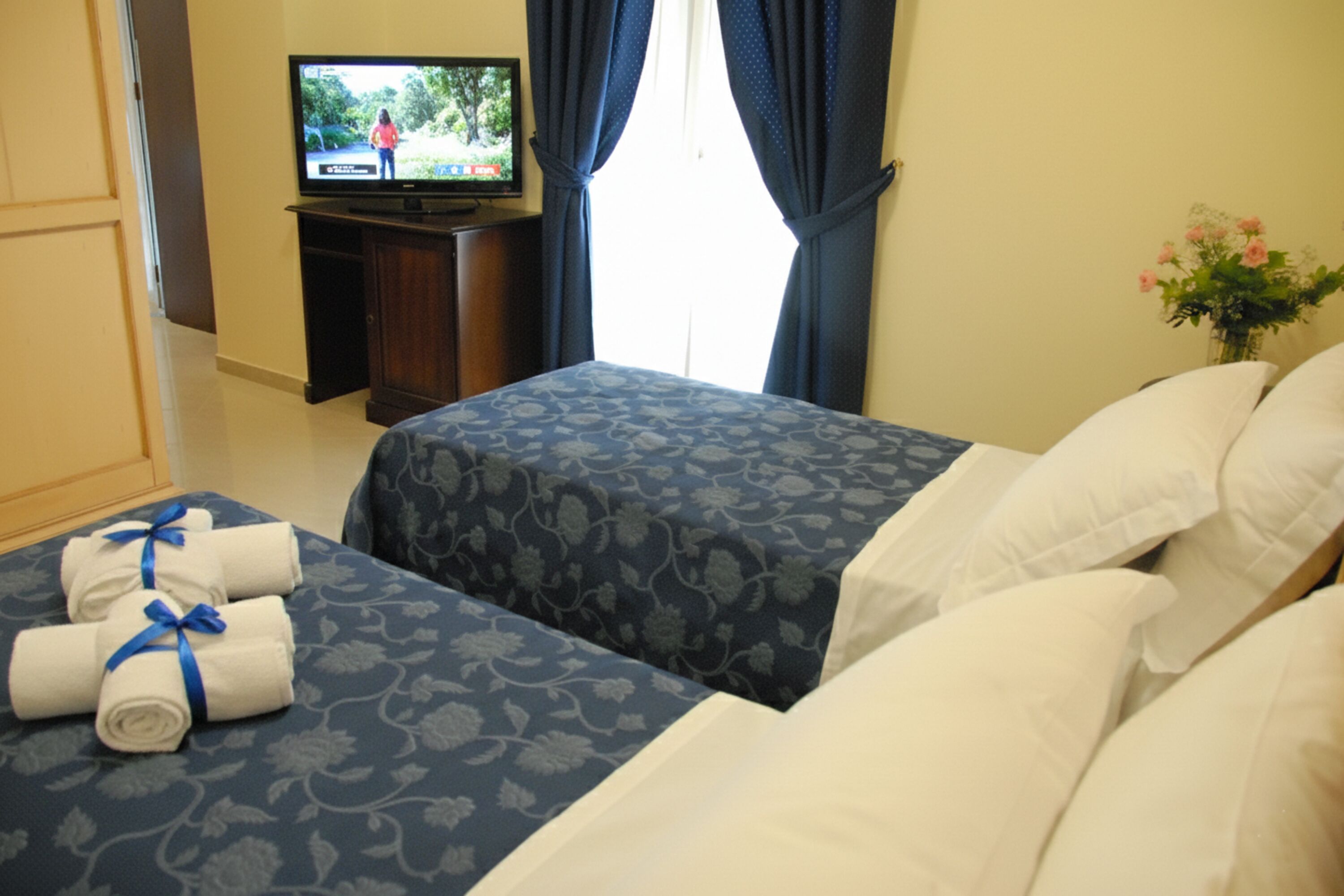A hotel room with two beds, a TV on a stand, a vase with flowers, and towels on the bed.