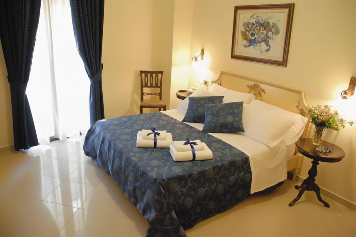 A neatly made bed with a blue bedspread and pillows, a bedside table with a lamp, a chair, and a painting on the wall.
