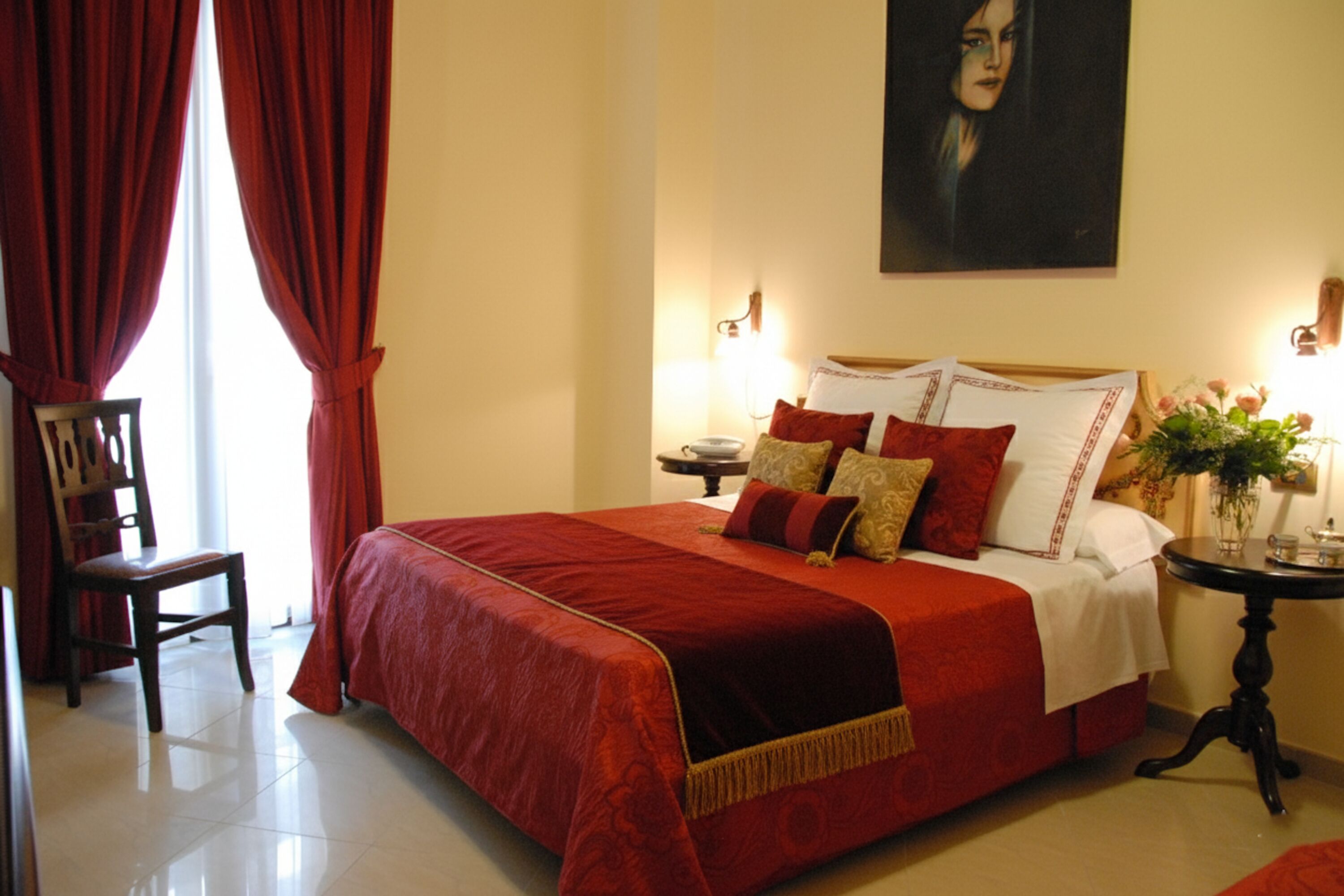 A neatly made bed with a red bedspread and pillows, a wooden chair, a bedside table with a vase of flowers, and a painting on the wall.