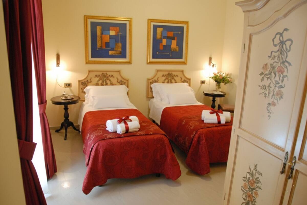 A hotel room with two beds, each with a red bedspread and white pillows. There are two framed artworks on the wall, a small table with a lamp, and a cabinet with floral designs.