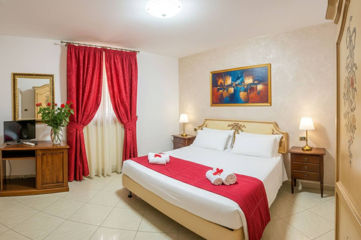 A hotel room with a bed, two bedside tables with lamps, a desk with a TV, a mirror, and red curtains.