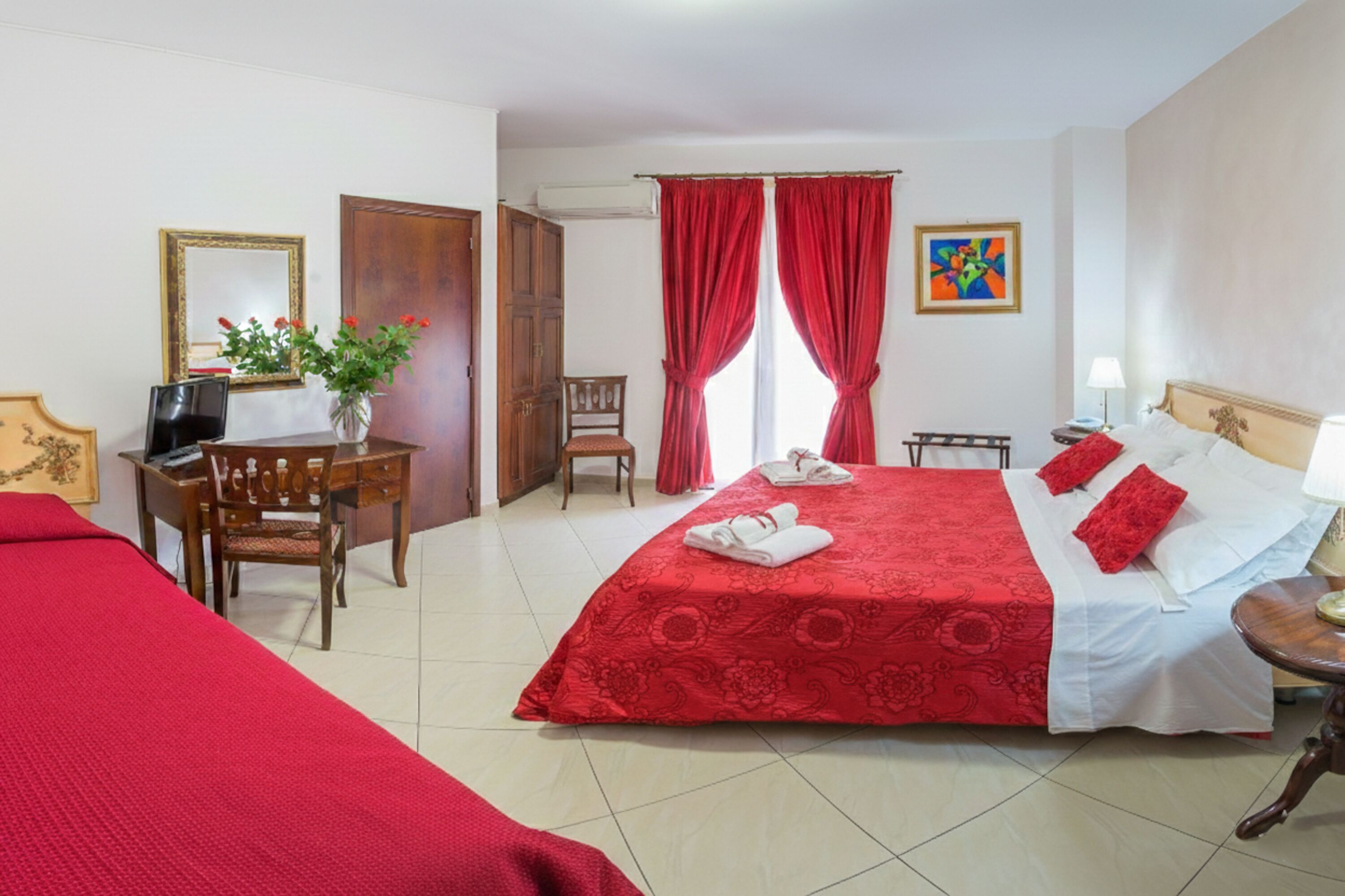 A hotel room with a bed, a desk, a chair, a TV, and a window with red curtains.