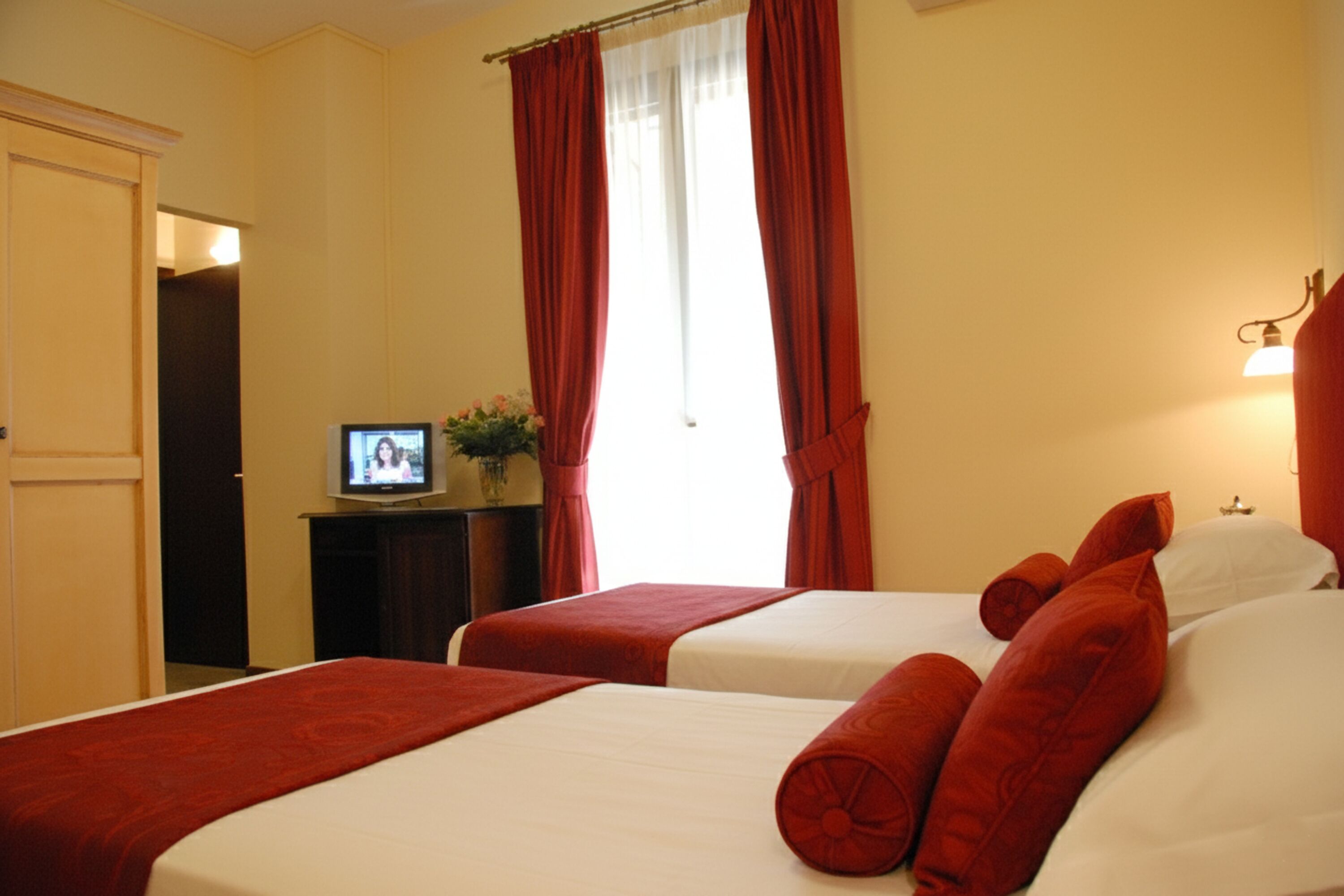 Een hotelkamer met twee bedden, rode gordijnen, een televisie op een tafeltje en een lamp aan de muur.