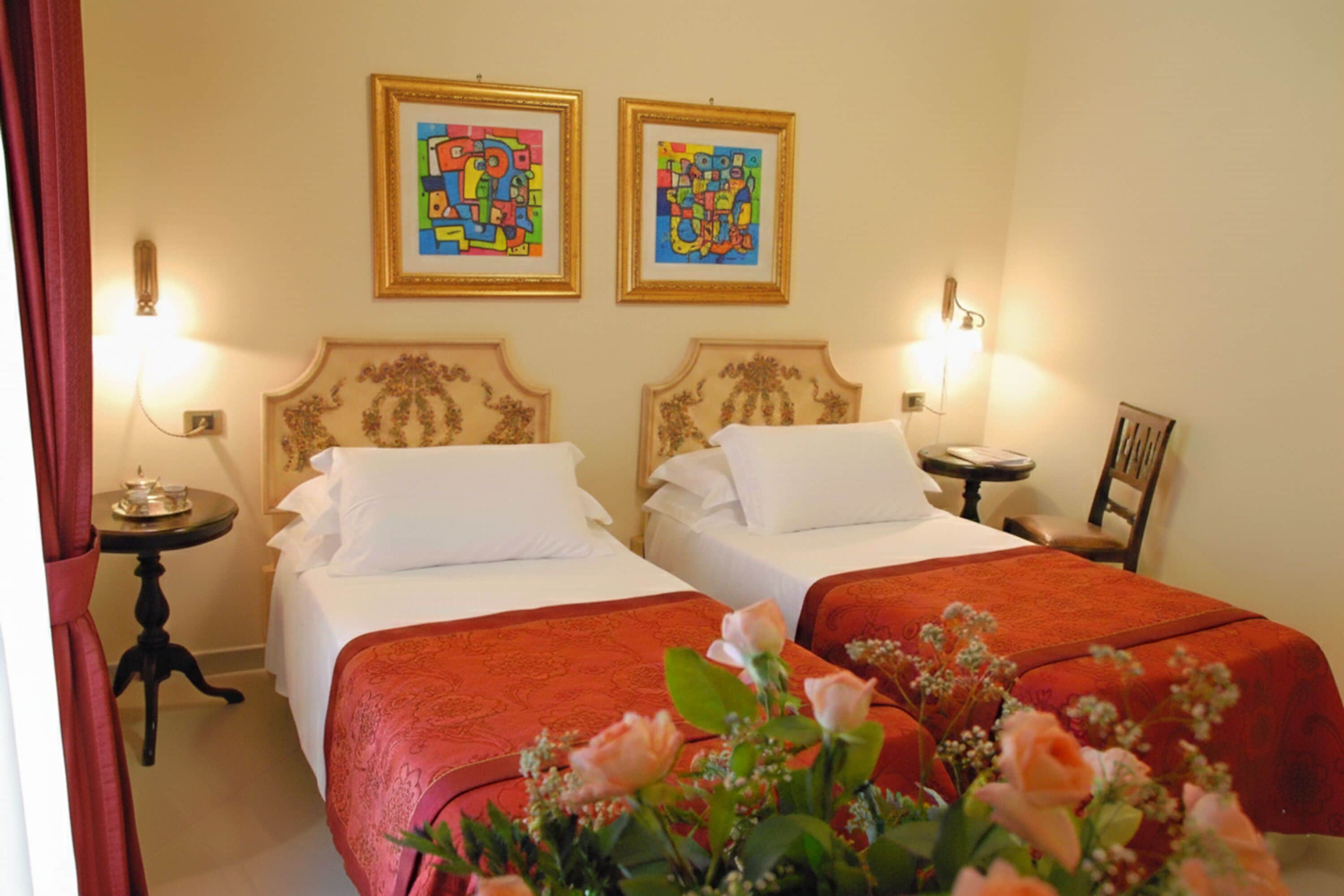 Une chambre d’hôtel avec deux lits, chacun recouvert d’une couverture rouge, agrémenté d’oreillers blancs et doté d’une tête de lit ornée de motifs floraux. Deux œuvres d’art encadrées sont accrochées au mur, au-dessus des lits.