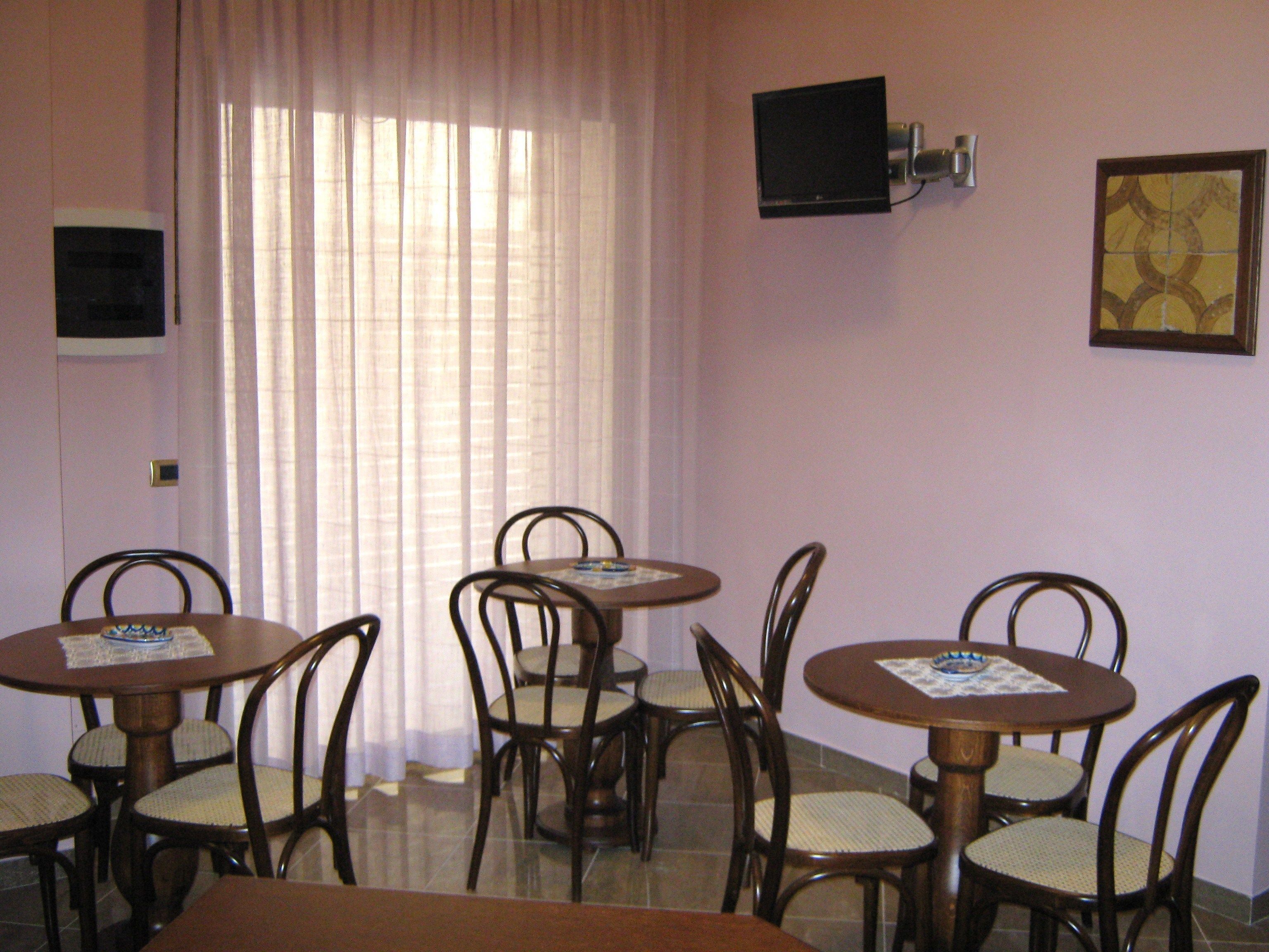 Una habitación con cuatro mesas y sillas de madera, un televisor montado en la pared y una ventana con cortinas traslúcidas.