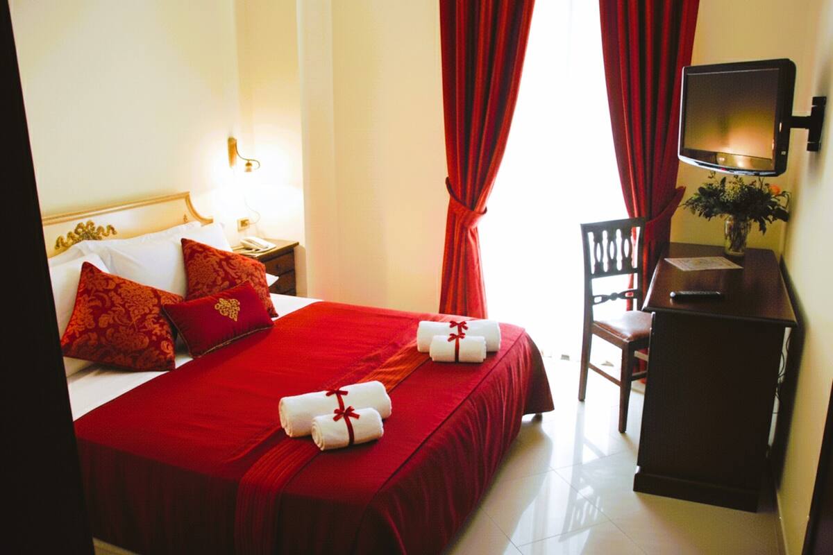 A hotel room with a large bed, red bedding, a desk with a television, and a chair.