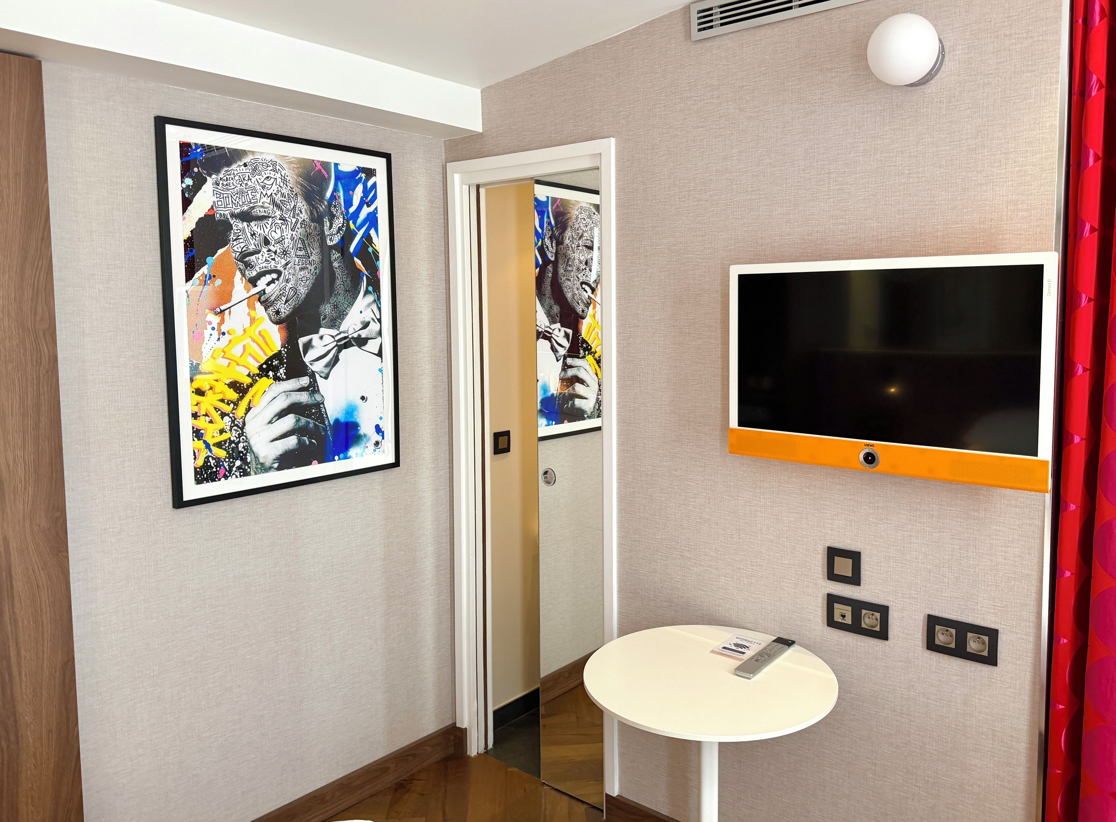 A hotel room with a large framed artwork, a flat-screen TV, a small table with a remote, and a door leading to another room.