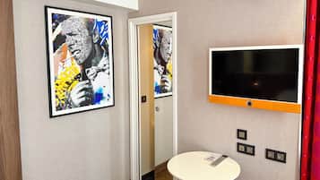 A hotel room with a large framed artwork, a flat-screen TV, a small table with a remote, and a door leading to another room.