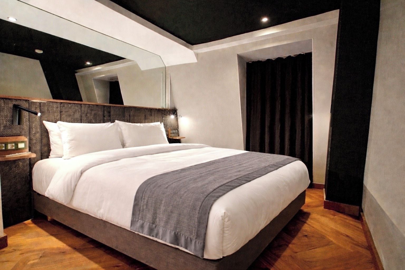 A modern hotel room with a large bed, a bedside table, a mirror, and a dark curtain.