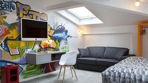 A bedroom with a skylight, a TV, a grey sofa, a white chair, and a bed with a grey blanket. - Hôtel Georgette (Paris)