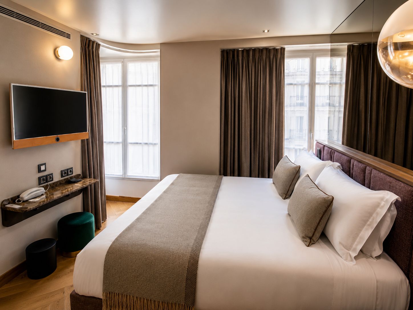 A hotel room with a large bed, a mounted TV, a bedside table with a phone, and a view of the city through the window.