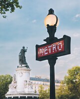 Una señal de calle con la palabra 'METRO', un farol y una estatua al fondo.