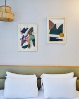 Ένα δωμάτιο ξενοδοχείου με ένα κρεβάτι, δύο μαξιλάρια, ένα προσκέφαλο και δύο αφηρημένα έργα τέχνης σε κορνίζες τοποθετημένα στον τοίχο.