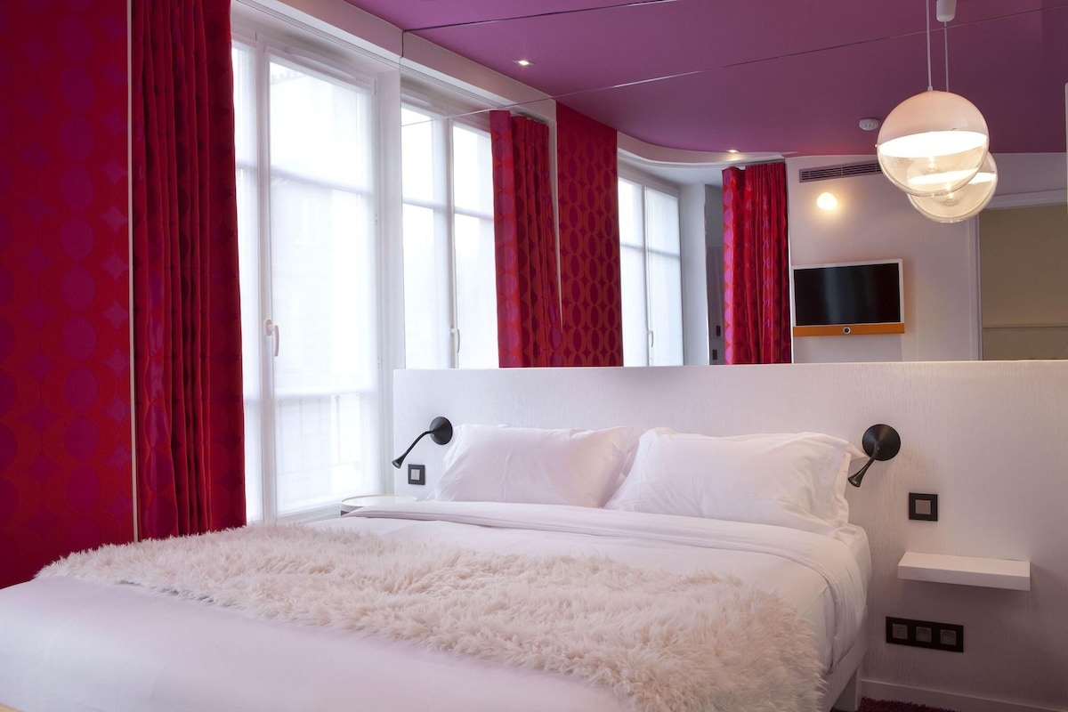 A modern bedroom with a large bed, a flat-screen TV mounted on the wall, and a ceiling light fixture.