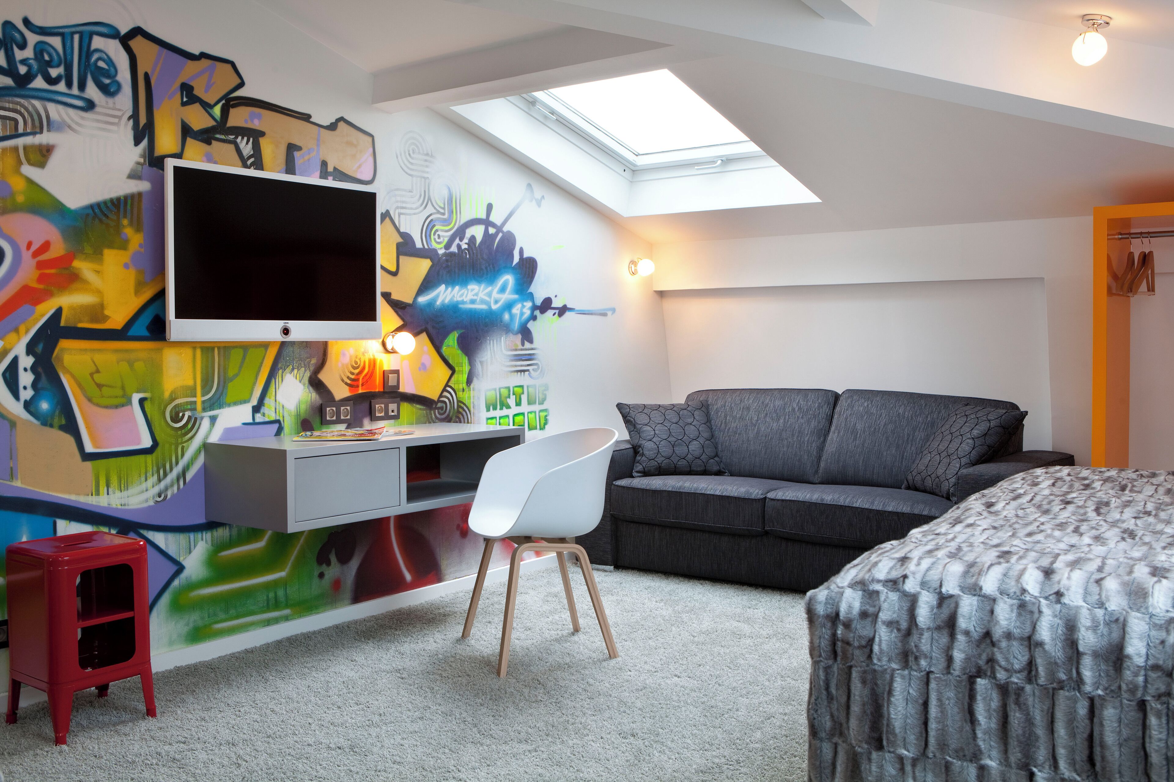 Une pièce avec une télévision, un bureau, un canapé gris, un tabouret rouge et un mur décoré de graffitis.