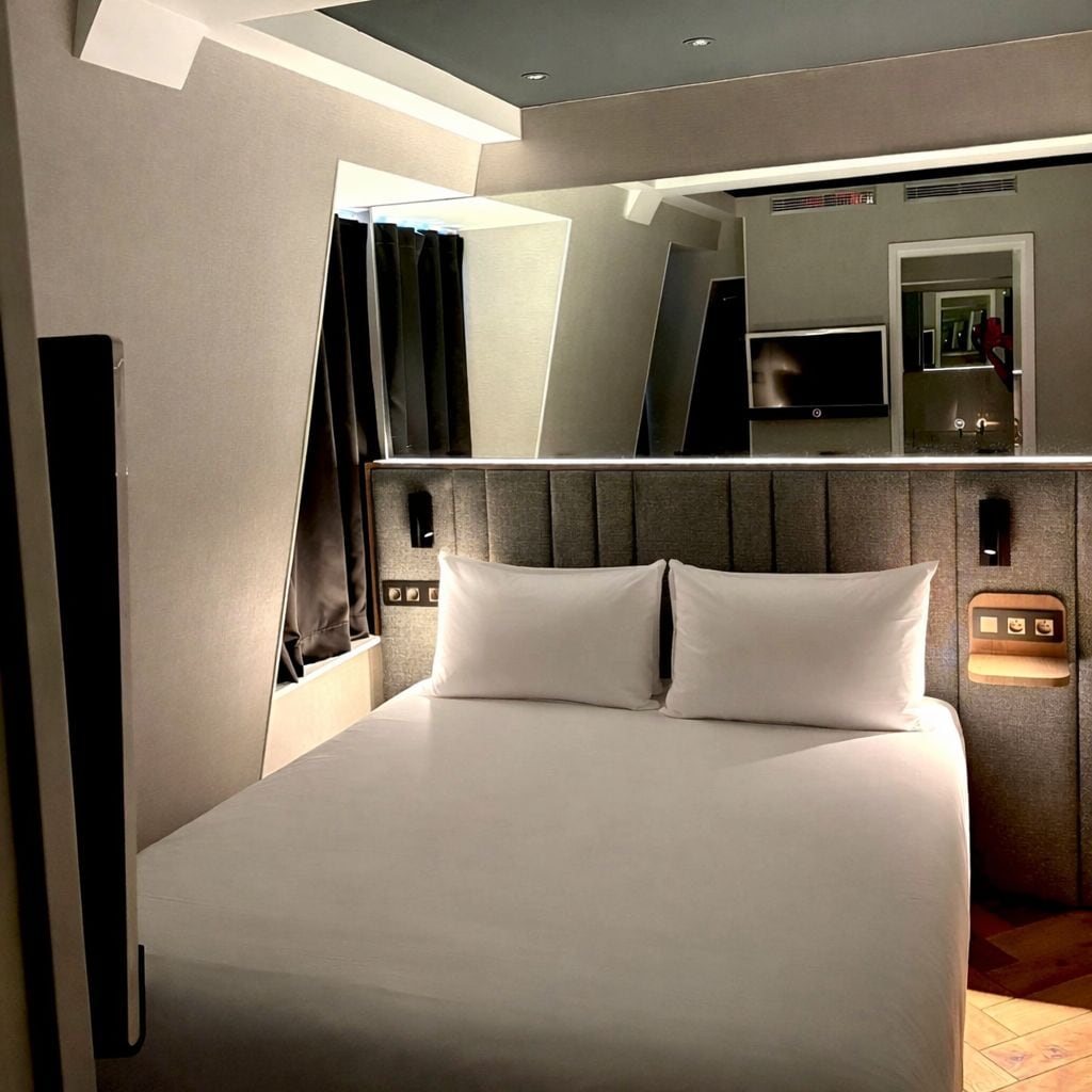 A modern hotel room with a large bed, a flat-screen TV, and a mirror above the bed.
