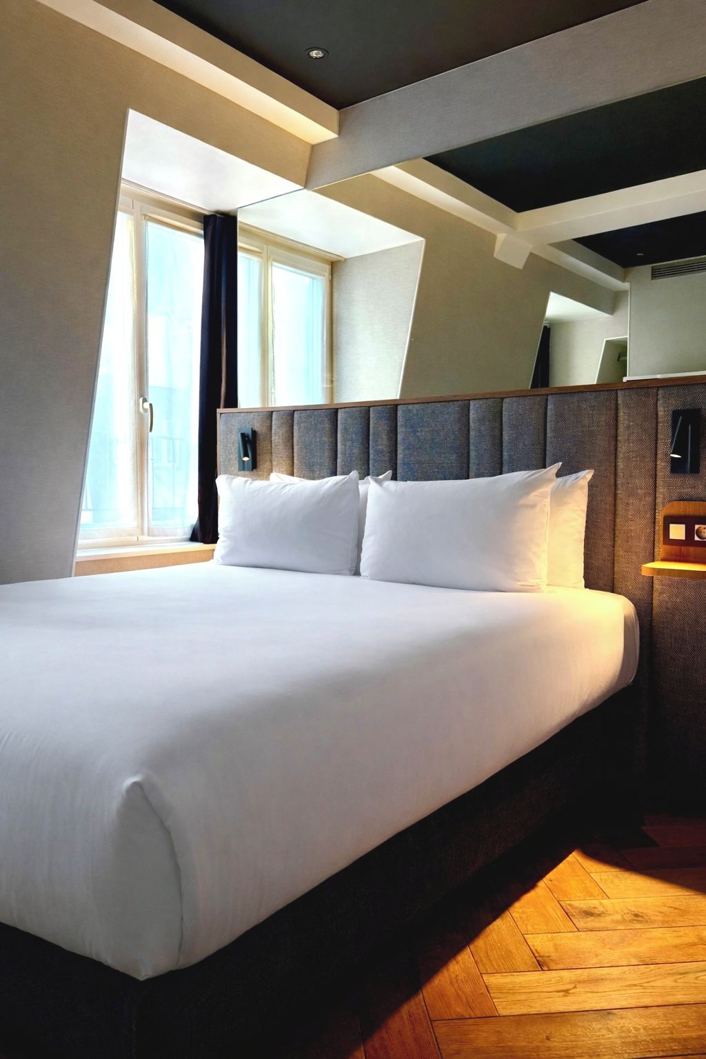 A modern hotel room with a large bed, a headboard with built-in lighting, a wooden floor, and a large window with curtains.