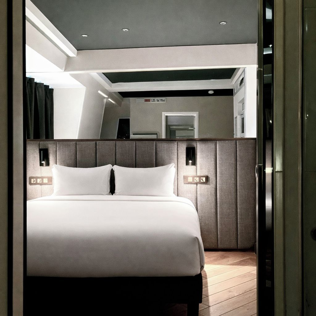 A modern hotel room with a large bed, a headboard with built-in lighting, and a mirror above the bed.