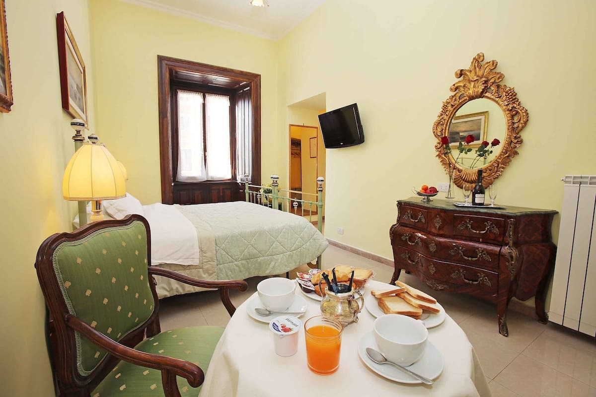 A hotel room with a bed, a dining table with breakfast items, a green armchair, a wooden dresser, and a mirror.