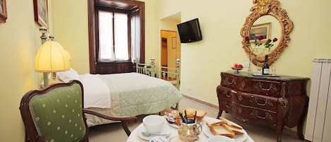 Habitación de hotel con una cama, una mesa de comedor con alimentos para el desayuno, un sillón verde, un aparador de madera y un espejo.