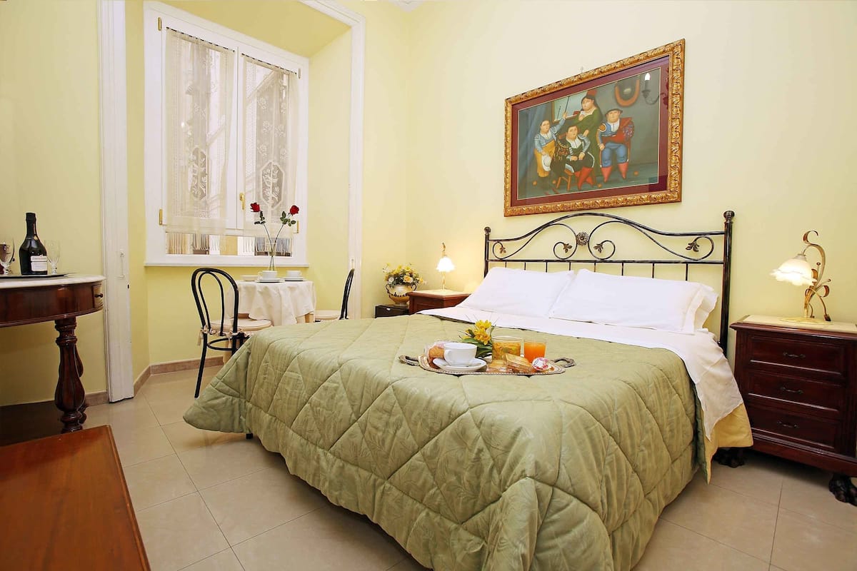 A neatly arranged bedroom with a green quilted bedspread, a painting on the wall, and a breakfast tray on the bed.