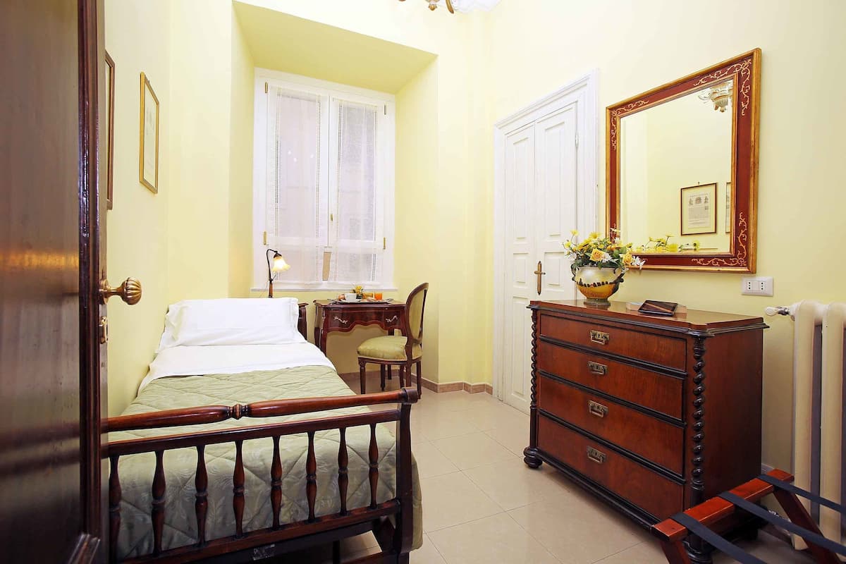 A bedroom with a wooden bed, a dresser, a chair, a window with white curtains, and a mirror with a decorative frame.