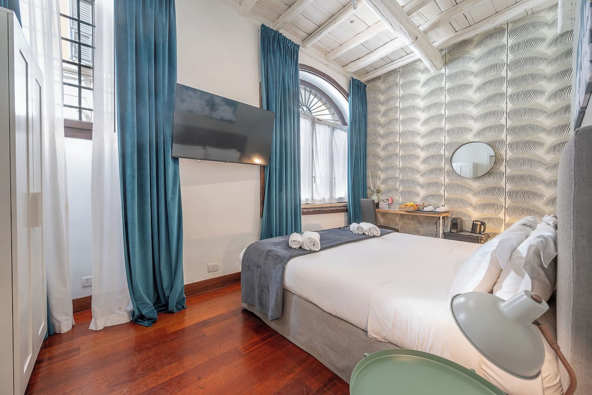 Une chambre d’hôtel avec un lit, une télévision, une fenêtre avec des rideaux, un plancher en bois et un mur orné d’un motif.