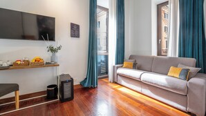 Un salon comprenant un canapé, un téléviseur à écran plat, une petite table avec des fruits et un vase, ainsi qu’une porte donnant sur un balcon.