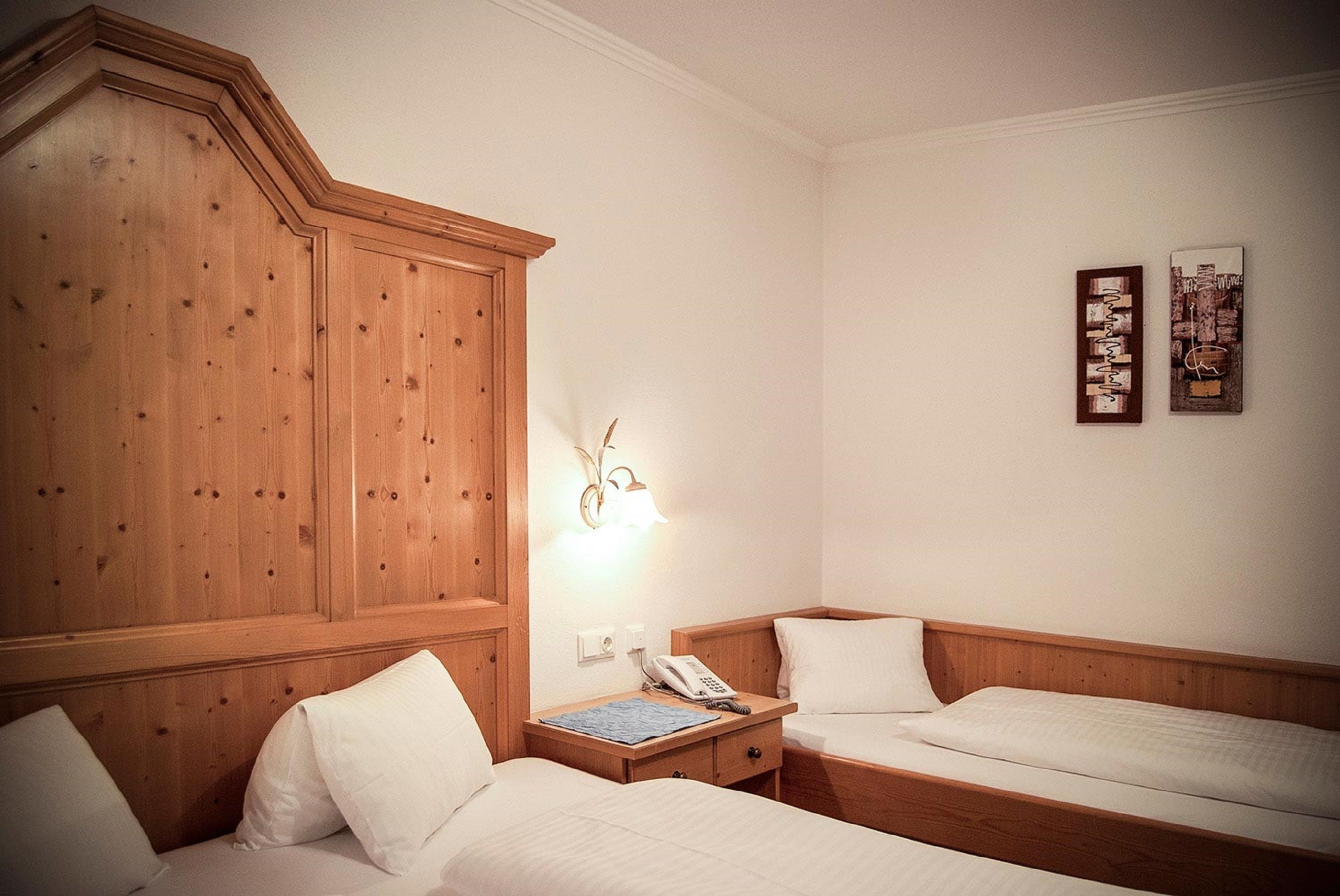A hotel room with two single beds, a wooden wardrobe, a bedside table with a lamp, and a wall-mounted phone.