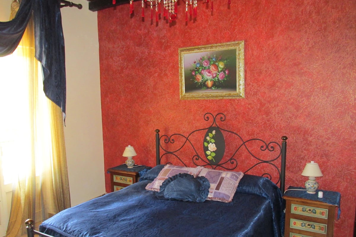Un dormitorio con cama, dos mesitas de noche, una lámpara en cada una, un cuadro en la pared y una ventana con cortinas.