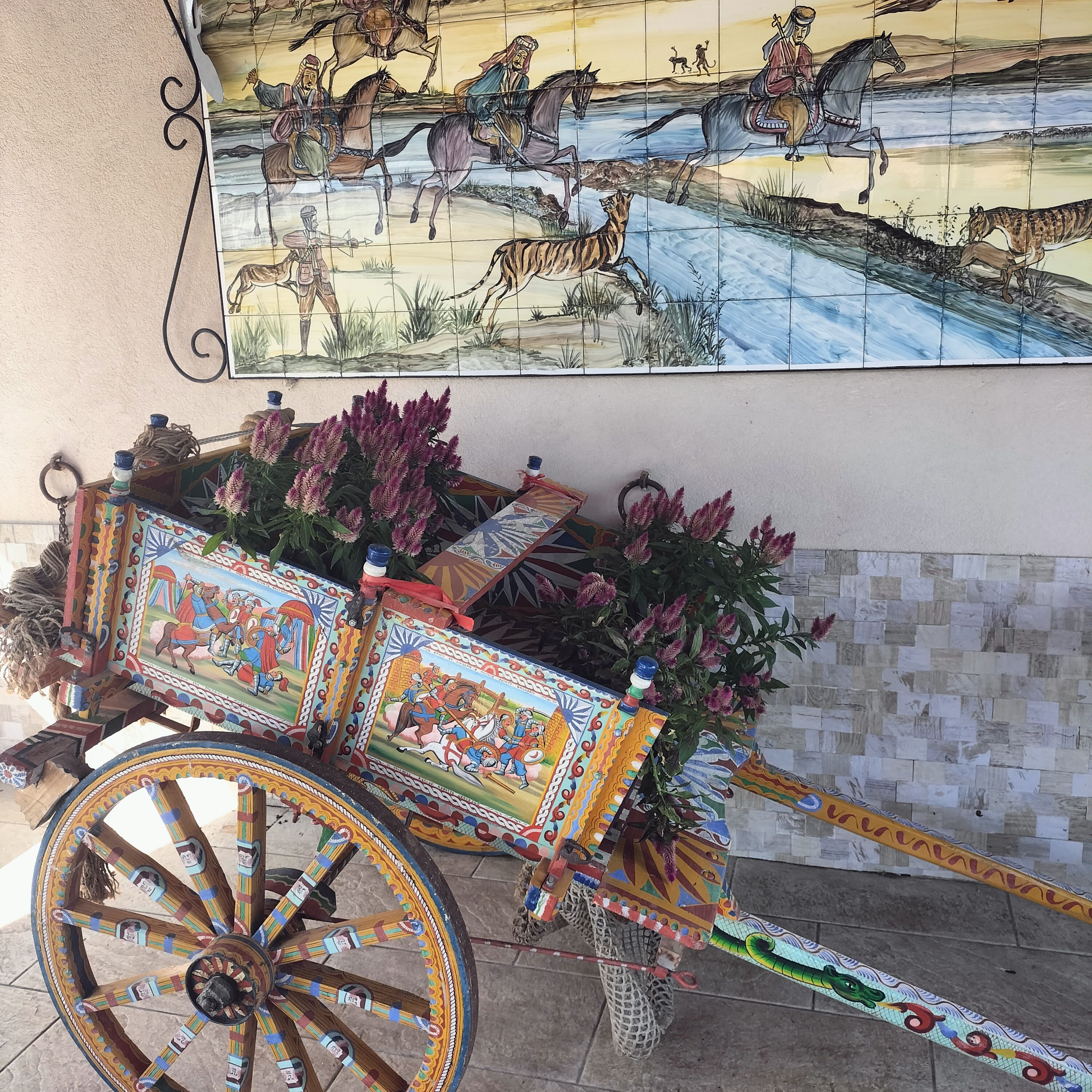 Un chariot décoratif orné de motifs complexes, une fresque représentant une scène historique avec des chevaux et des cavaliers, et un mur recouvert de carreaux formant un motif géométrique.