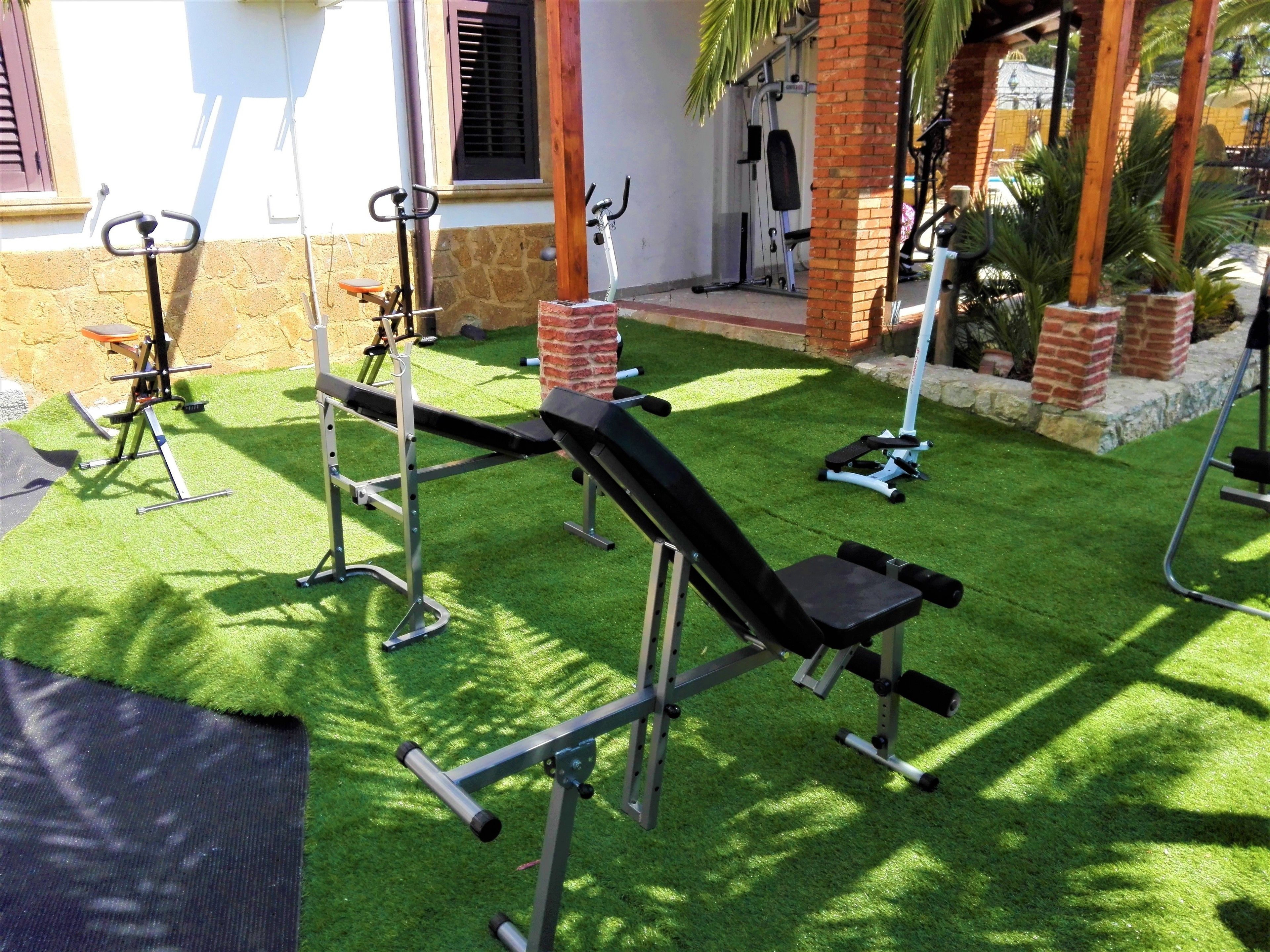An outdoor gym area with various exercise equipment, including a bench press, a rowing machine, and a leg press, set against a backdrop of a building with a brick facade and a small garden with palm trees.